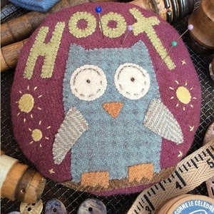 May include: A round, burgundy pincushion with a felt owl design. The owl is gray with orange feet and a beak. The pincushion is decorated with the word "HOOT" in yellow felt letters and yellow felt suns.