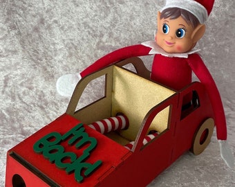 May include: A red wooden toy car with a green "I'm back!" sign on the hood. A Christmas elf doll is sitting in the car with its legs sticking out.