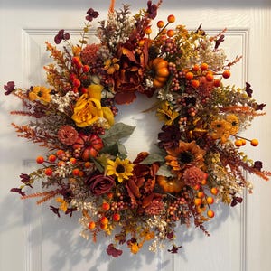 May include: A circular autumn wreath featuring a dense arrangement of artificial flowers, including yellow roses, sunflowers, and deep red blossoms. The design incorporates small orange pumpkins, clusters of red and orange berries, and dried foliage.