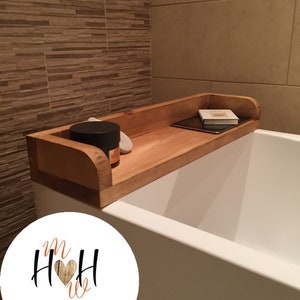 May include: Wooden bath tray with rounded edges, designed to fit across a bathtub. The tray has a small book and a tablet on it. The tray is made of natural wood and has a rustic finish. The tray is labelled with the text "Handmade Wooden Home".