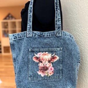 May include: A denim tote bag with a light blue wash. The bag features a front pocket with a watercolor image of a Highland cow adorned with pink flowers. The bag has two denim handles.