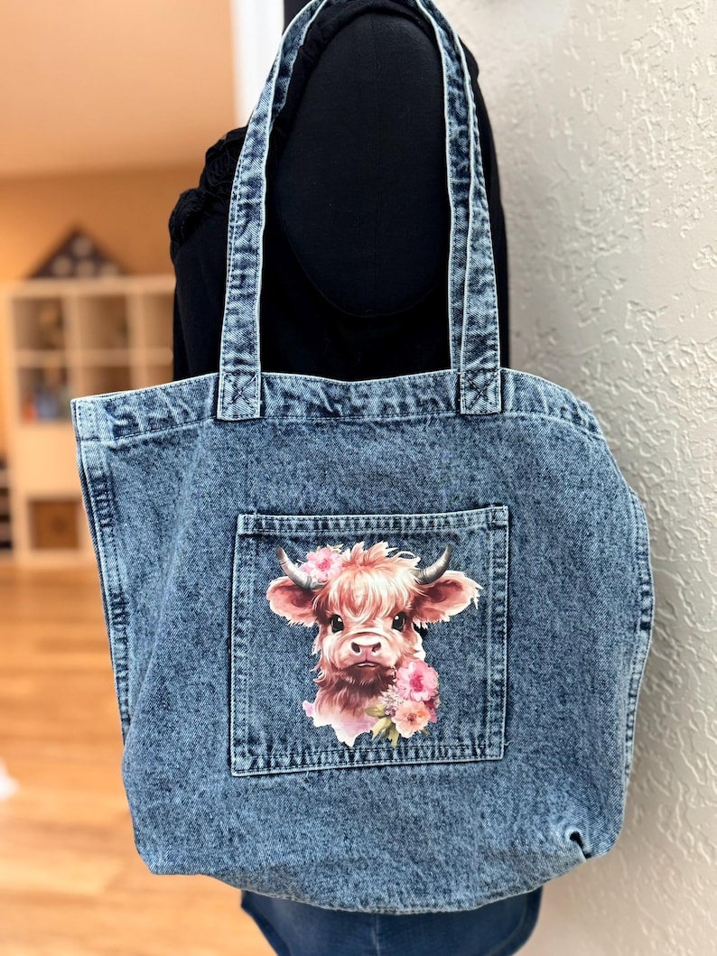 May include: A denim tote bag with a light blue wash. The bag features a front pocket with a watercolor image of a Highland cow adorned with pink flowers. The bag has two denim handles.