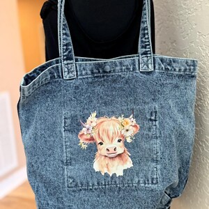 May include: Denim tote bag with a light blue wash. The bag features a pocket with a graphic of a Highland cow wearing a floral crown. The bag has two denim handles.