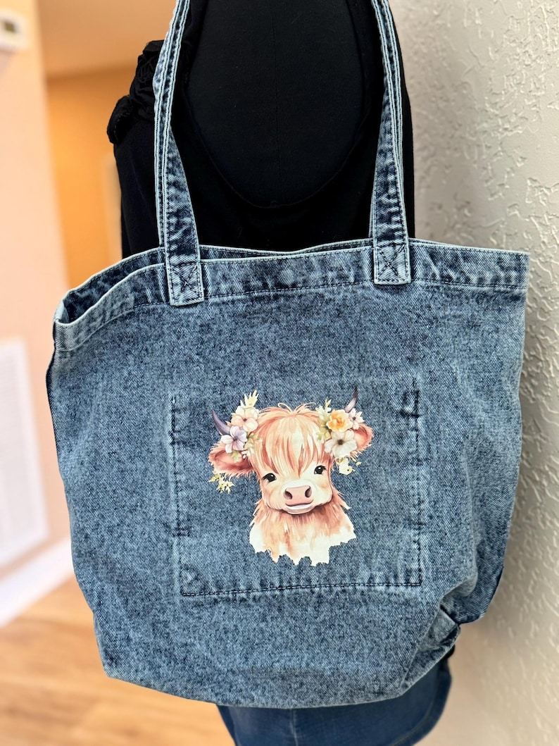 May include: Denim tote bag with a light blue wash. The bag features a pocket with a graphic of a Highland cow wearing a floral crown. The bag has two denim handles.