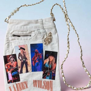 May include: A white denim crossbody bag featuring four rectangular photo prints of a musician playing guitar and wearing a cowboy hat. The bag has a gold chain strap with white woven accents and pearl details, a small zipper pocket, and the text LAINEY WILSON.