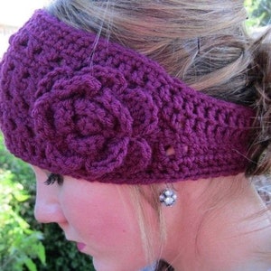 May include: A purple crocheted headband with a large flower design. The headband is being worn by a person with blonde hair.