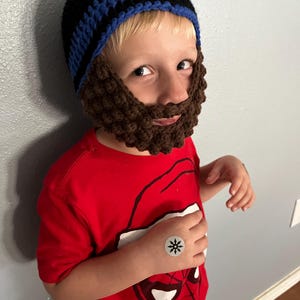 May include: A young child wearing a blue and black striped crocheted hat with a brown crocheted beard attached. The child is wearing a red t-shirt with a Spiderman graphic and a black star sticker on their wrist.