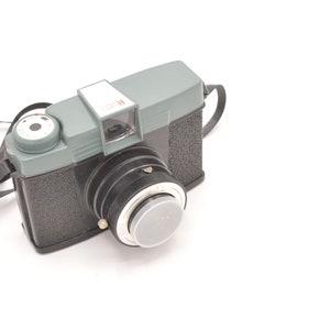 Peut inclure: Un appareil photo en plastique noir et vert avec une sangle noire. L'appareil photo a un cache d'objectif et une étiquette rouge "K-25" sur le dessus.