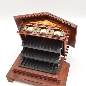 May include: A wooden chalet-style cigar humidor featuring a dark brown finish, a gabled roof with scalloped edges, and three black tiered trays with molded grooves for cigar storage. The facade includes three small windows with green shutters.