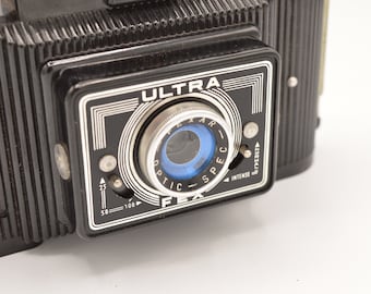 May include: A black vintage camera with a silver lens and the word "ULTRA" printed on the front. The camera has a dial for adjusting the aperture and a dial for adjusting the shutter speed.