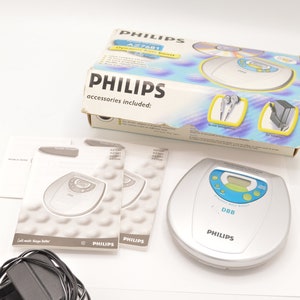 May include: A silver Philips portable CD player with a blue display and the text "DYNAMIC BASS BOOST" and "DBB". The player is on a white surface with a black power adapter, a pair of headphones, and two instruction manuals.