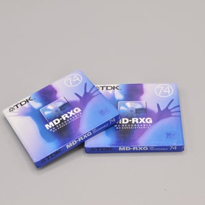 Könnte beinhalten: Drei blaue und violette TDK MD-RXG 74 Minuten Mini-Disc-Aufnahmehüllen mit einer silbernen Scheibe und einer Silhouette einer Hand im Hintergrund.