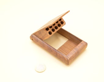 Puede incluir: Una caja de madera con una tapa con bisagras que se abre para revelar una fila de pequeños agujeros redondos. La caja está hecha de madera oscura con un patrón de veta natural.