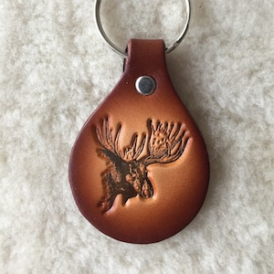 May include: A brown leather keychain with a moose head design. The moose is carved into the leather and has a detailed face and antlers.