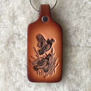 May include: A brown leather keychain with a key ring and a design of two quail in a grassy field.