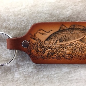 May include: A brown leather keychain with a silver key ring. The keychain features an engraved image of a redfish in a marsh setting.