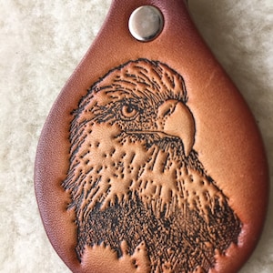 May include: A brown leather keychain with a detailed embossed image of a bald eagle's head. The keychain has a silver metal ring and a silver metal button.