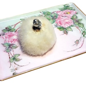 May include: A white fluffy powder puff with a silver top sits on a porcelain tray with a pink and green floral design.