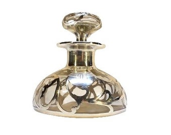 May include: A clear glass bottle with a silver filigree design and a gold metal stopper. The bottle has a round body and a short neck.