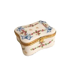 Puede incluir: Una caja de porcelana blanca para alhajas con un diseño floral y una cinta azul. La caja tiene una tapa con bisagras y un cierre de metal dorado.