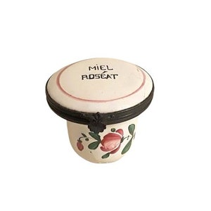 May include: A small white ceramic jar with a lid. The lid has a black metal rim and the words "MIEL ROSEAT" written on it. The jar is decorated with pink roses and green leaves.