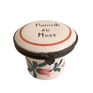 Puede incluir: Un tarro de cerámica blanca con una tapa con bisagras. La tapa está decorada con una banda roja y las palabras "Pommade au Muse". El tarro está decorado con hojas verdes y flores rosas.