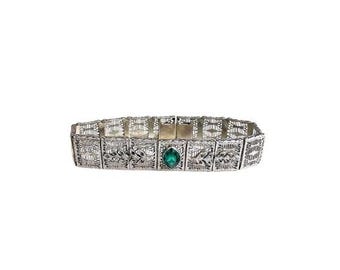 May include: A silver filigree bracelet with an emerald-green gemstone at its centre. The bracelet has an openwork design with floral and geometric patterns. The clasp is visible. A piece of jewellery.