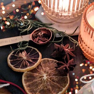 May include: A copper measuring spoon filled with cloves and star anise, next to dried orange slices and rosemary sprigs. Two lit candle holders are in the background, with bokeh lights.