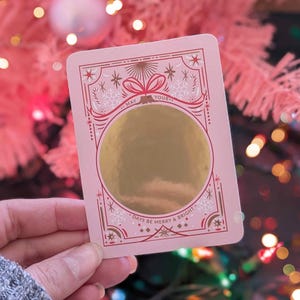 May include: A pink holiday scratch-off card with gold foil and decorative snowflake and bow accents. The card reads "May Your Days Be Merry & Bright" in a festive font. The card is rectangular with rounded corners.
