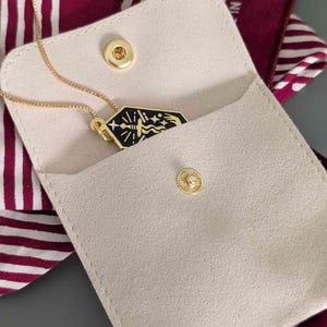 May include: A gold-toned necklace with a black pendant featuring a stylized design, partially visible inside a beige suede pouch. The pouch has a gold snap closure and is set against a patterned burgundy background.