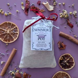 May include: A beige fabric bag with a tag reading "SIMMER POT SELF-LOVE" tied with a red ribbon. The image shows dried orange slices, cinnamon sticks, and dried flowers scattered on a purple surface. The tag lists ingredients like jasmine, apple, and lavender.