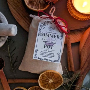 May include: A beige fabric bag with a tag that reads "SIMMER POT SELF-LOVE". The tag lists a blend of dried jasmine, rose, chamomile, lemon, orange, apple, and lavender. The bag is tied with a red ribbon. Dried orange slices and a lit candle are in the background.