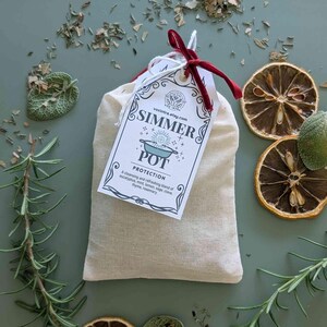 May include: A beige muslin bag with a tag that reads "SIMMER POT PROTECTION". The tag is tied with a red ribbon. The bag is surrounded by dried lemon slices, rosemary sprigs, and other herbs on a green surface.