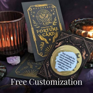 May include: Two black rectangular scratch-off fortune cards with gold foil accents and illustrations of hands and eyes. One card features the text "SCRATCH OFF FORTUNE CARD" and the other displays the text "Your strength lies not in avoiding pain but in embracing it with courage; in surprising yourself with your own resilience."