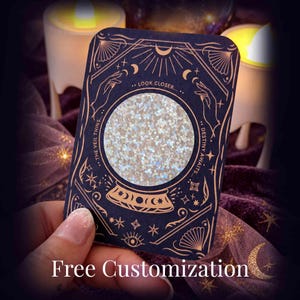 May include: A rectangular dark blue card featuring a central circular cutout filled with iridescent white glitter. The card is decorated with copper-colored line art of hands, stars, and moons, with the text "THE VEIL THINS... LOOK CLOSER... DESTINY AWAITS..." printed around the circle.