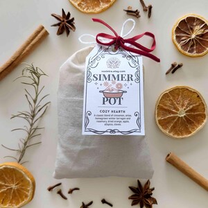 May include: A beige fabric bag with a tag reading "SIMMER POT COZY HEARTH", tied with a red ribbon. The image features dried orange slices, cinnamon sticks, star anise, and cloves, creating a warm, inviting aesthetic.