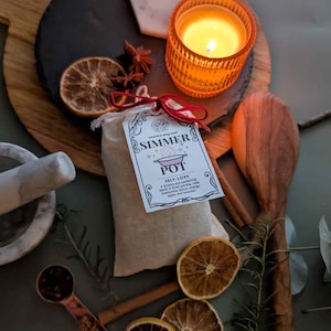 May include: A still-life arrangement featuring a lit orange candle in a glass holder, a small bag labeled "SIMMER POT SELF-LOVE", dried orange slices, and a wooden spoon. The scene includes a mortar and pestle, star anise, and cinnamon sticks.