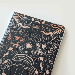 May include: A spiral-bound notebook with a black cover featuring a rose gold celestial design. The design includes suns, moons, stars, snakes, birds, and other mystical elements. The notebook is open, revealing the white spiral binding.