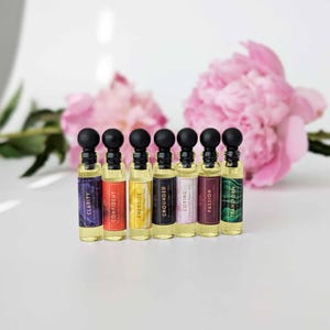 May include: A row of seven small perfume oil bottles with black caps and colourful labels. The labels read "Clarity", "Confident", "Energize", "Grounded", "Loving", "Passion", and "Transform". Pink flowers are in the background.