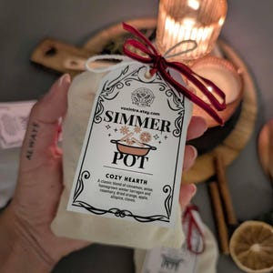 May include: An off-white bag with a tag that reads "SIMMER POT" with the text "COZY HEARTH". The tag is tied with a red ribbon. The bag contains a blend of cinnamon, anise, tarragon, rosemary, dried orange, apple, allspice, and cloves.