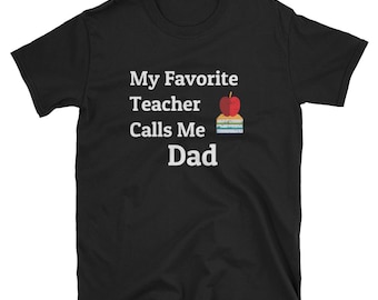 Op de afbeelding: Zwart T-shirt met witte tekst die "Mijn favoriete leraar noemt me papa" luidt. Een rode appel en een stapel boeken bevinden zich boven de tekst.
