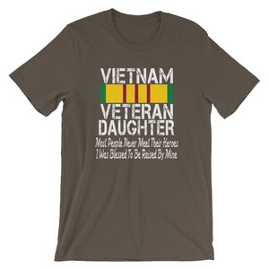 May include: A dark brown t-shirt with a white graphic print that says "VIETNAM VETERAN DAUGHTER" with a green, red, and yellow ribbon above the text. The text below the ribbon says "Most People Never Meet Their Heroes I Was Blessed To Be Raised By Mine."