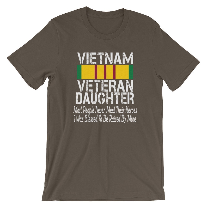 May include: A dark brown t-shirt with a white graphic print that says "VIETNAM VETERAN DAUGHTER" with a green, red, and yellow ribbon above the text. The text below the ribbon says "Most People Never Meet Their Heroes I Was Blessed To Be Raised By Mine."
