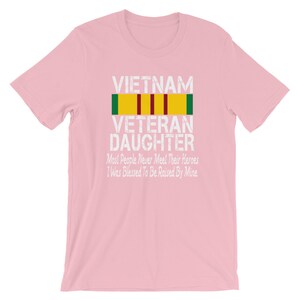 May include: Pink t-shirt with a white graphic that says "VIETNAM VETERAN DAUGHTER" with a yellow, red, and green ribbon above the text. The graphic also says "Most People Never Meet Their Heroes I Was Blessed To Be Raised By Mine".