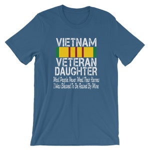 May include: A blue t-shirt with a white distressed graphic that reads "VIETNAM VETERAN DAUGHTER" with a green, yellow, and red ribbon above the text. The text below the ribbon reads "Most People Never Meet Their Heroes I Was Blessed To Be Raised By Mine."