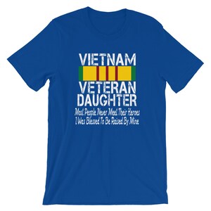 May include: Blue t-shirt with a white graphic that says "VIETNAM VETERAN DAUGHTER" with a yellow and green ribbon above the text. The text below the ribbon says "Most People Never Meet Their Heroes I Was Blessed To Be Raised By Mine".
