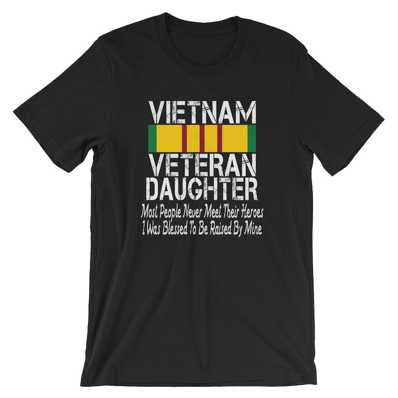 May include: Black t-shirt with white text that reads "VIETNAM VETERAN DAUGHTER" with a yellow, green, and red ribbon above the text. The text below the ribbon reads "Most People Never Meet Their Heroes I Was Blessed To Be Raised By Mine."