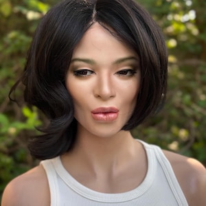 May include: A mannequin head featuring a short, dark brown bob hairstyle with a center part and soft waves. The mannequin has a realistic facial structure with defined eyebrows, dark eyeliner, and glossy lips, wearing a simple white ribbed tank top.