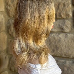 May include: A blonde wig with a layered style, featuring a blend of light and dark tones. The hair is styled with soft waves and a side part. The wig is worn over a white ribbed top, set against a stone wall background.