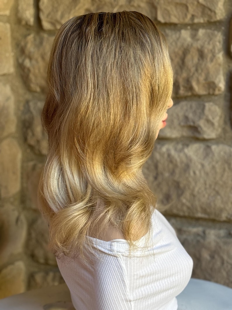 May include: A blonde wig with a layered style, featuring a blend of light and dark tones. The hair is styled with soft waves and a side part. The wig is worn over a white ribbed top, set against a stone wall background.